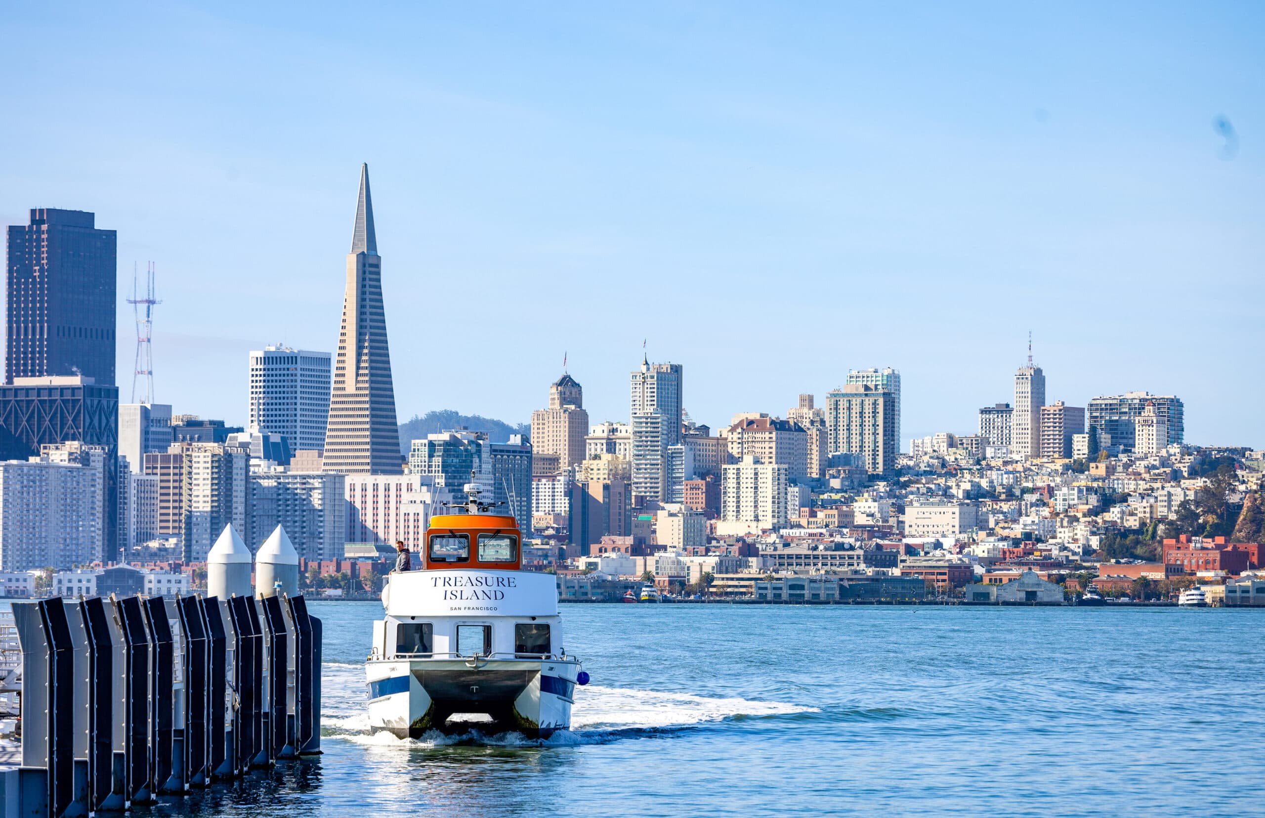 Treasure Island ferry cruising across San Francisco Bay with the downtown skyline and Transamerica Pyramid in the background on a clear blue day.