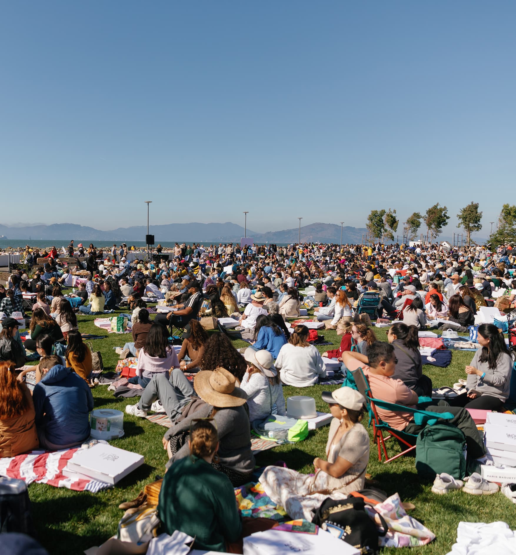 Large outdoor community gathering at a waterfront park in San Francisco, with hundreds of people seated on blankets and lawn chairs enjoying a sunny daytime event by the bay.