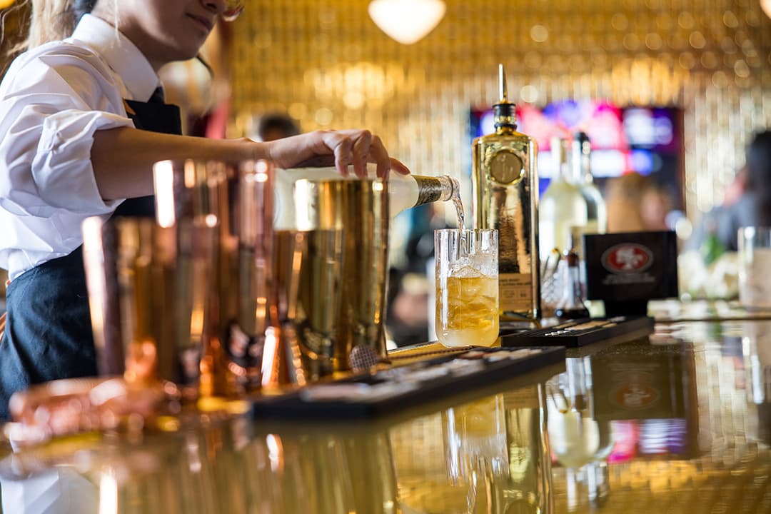 Bartender pouring a cocktail over ice at Gold Bar, with bottles, bar tools, and warm ambient lighting in the background.