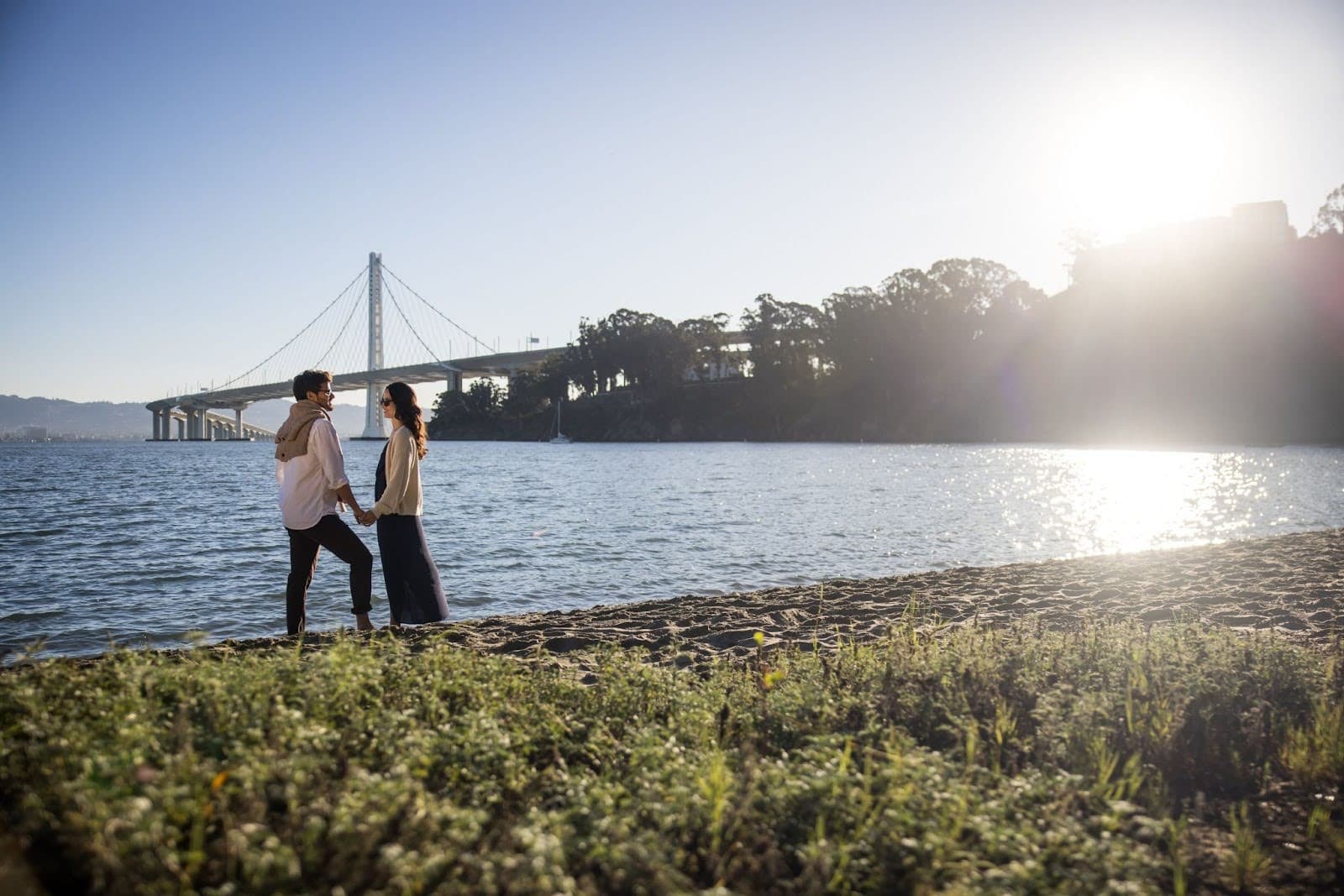 Couple walking along the Treasure Island shoreline at sunset with the Bay Bridge and San Francisco Bay in the background.