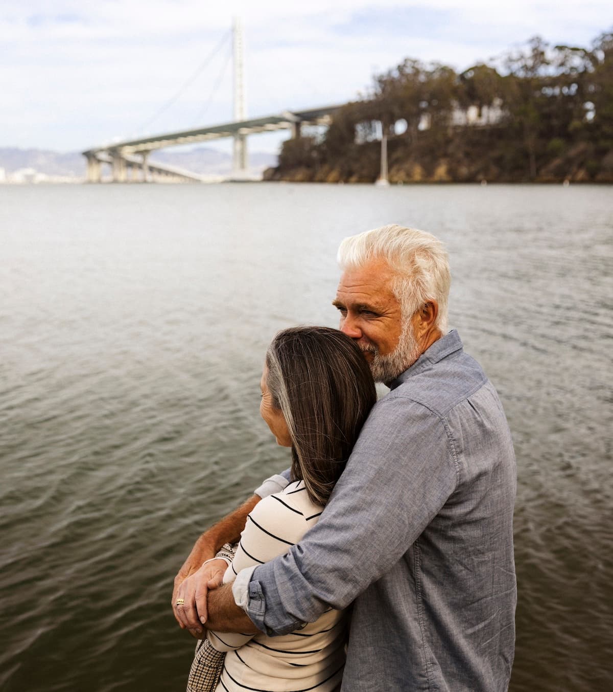 Older couple embracing by the waterfront with the San Francisco–Oakland Bay Bridge in the background on Treasure Island.