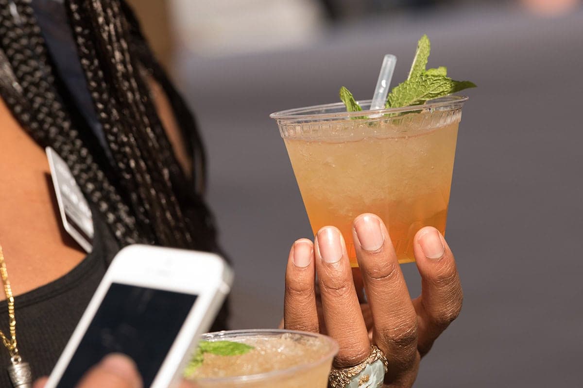 Close-up of a person holding a mint garnish cocktail in a clear plastic cup at an outdoor event while using a smartphone.