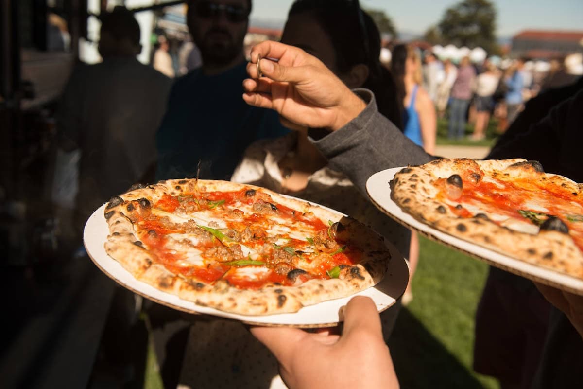 Fresh wood-fired Neapolitan pizzas being served on paper trays at an outdoor food festival with a crowd in the background.