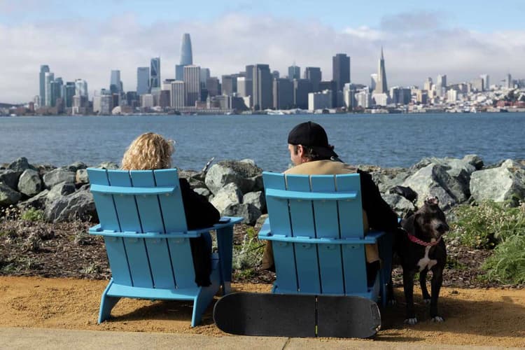 Two people sit in bright blue Adirondack chairs along the waterfront, looking across the bay at the San Francisco skyline, with a dog and skateboard beside them.
