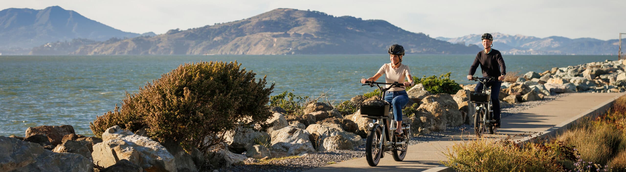 Two adults riding electric bikes along a paved waterfront trail by the San Francisco Bay, with rocky shoreline and rolling hills in the background on a clear, sunny day.