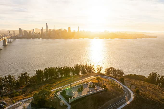 An elevated sunset view over a waterfront hillside park, with winding paths and a small sculptural feature in the foreground, and the San Francisco skyline glowing across the bay as the sun reflects off the water.