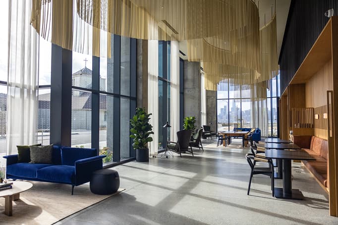 A modern, light-filled lounge space with floor-to-ceiling windows, blue velvet seating, wood accents, and city views in the distance.