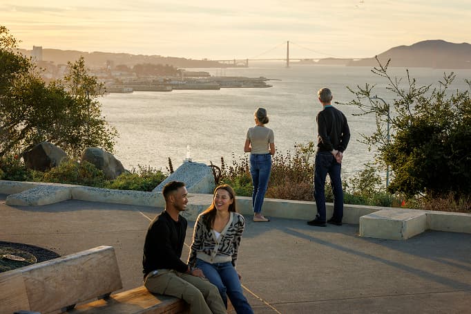 People enjoying a scenic overlook at sunset, with two individuals seated on a bench chatting while others stand at the railing taking in expansive views of the bay and distant bridges under warm golden light.
