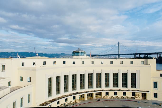 A historic Art Deco-style building with tall vertical windows and a curved façade, overlooking the bay, with the San Francisco–Oakland Bay Bridge visible in the background.