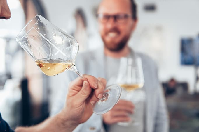 Two people tasting white wine indoors, with one person swirling a glass in the foreground while another smiles in the background holding their own glass, suggesting a relaxed winery or tasting room setting.