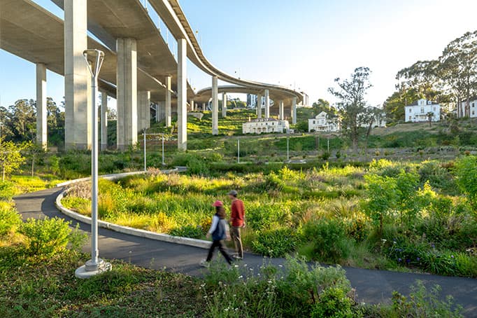 People walking along a paved path through Willow Bridge Park, with landscaped greenery and the Bay Bridge ramps rising overhead.