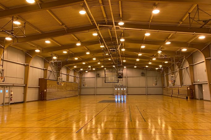 A spacious indoor gymnasium with polished wooden floors, basketball hoops, and bright overhead lighting, ready for sports or community activities.