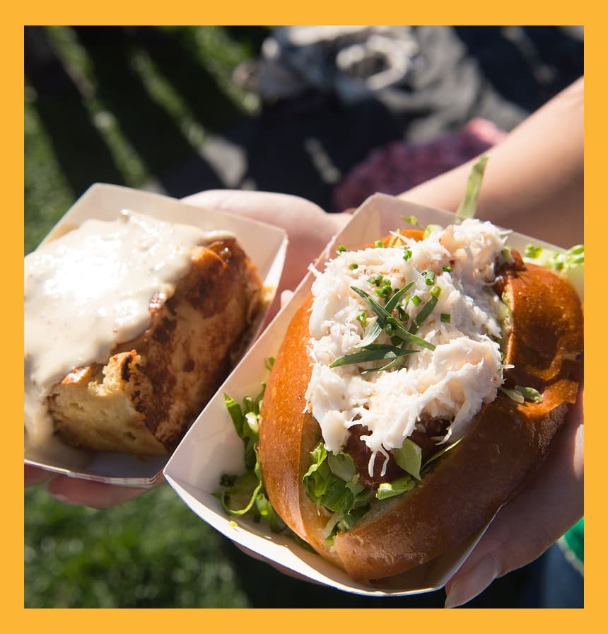 Close-up of two handheld food trays with a crab sandwich and a creamy baked dish