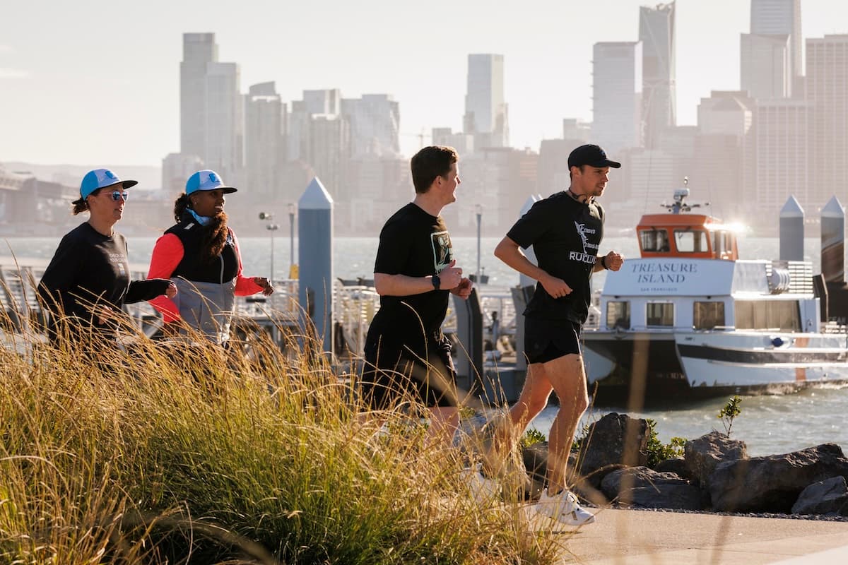 Four people jogging along a waterfront path with a city skyline and a docked boat in the background on a sunny day.