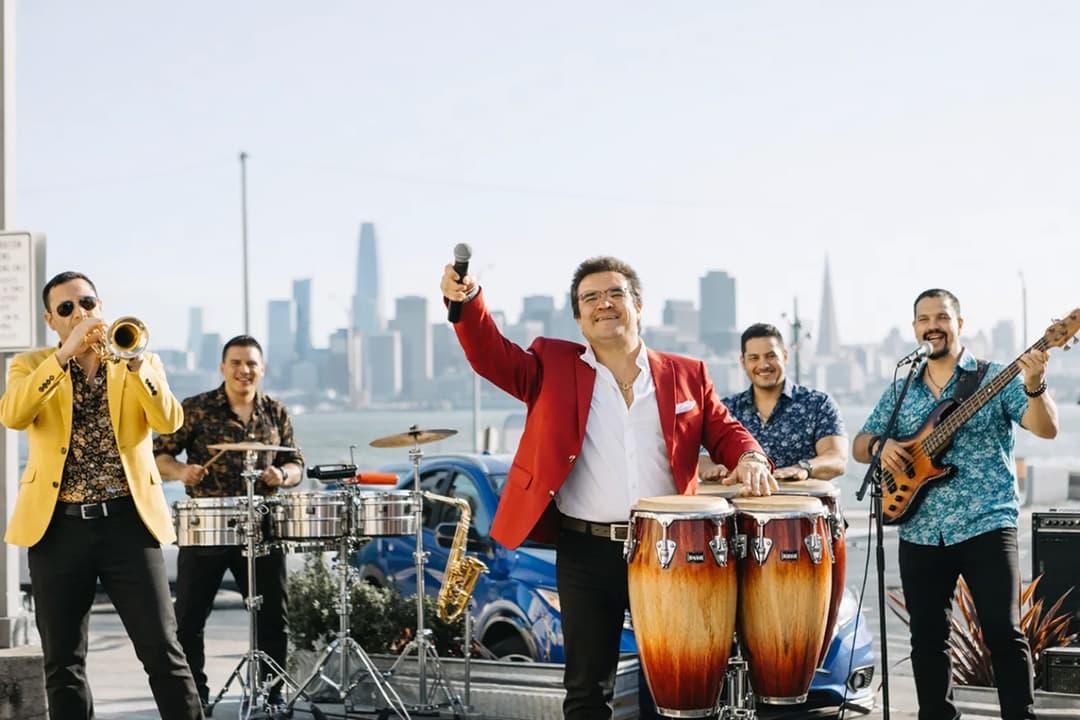 A lively Latin band performs outdoors on Treasure Island, with a singer in a red jacket playing congas and holding a microphone, surrounded by musicians on trumpet, drums, and bass, with the San Francisco skyline in the background.