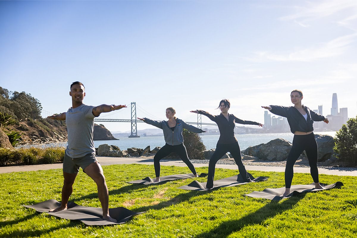 People practicing yoga on mats by the waterfront, with the Bay Bridge and San Francisco skyline in the background.