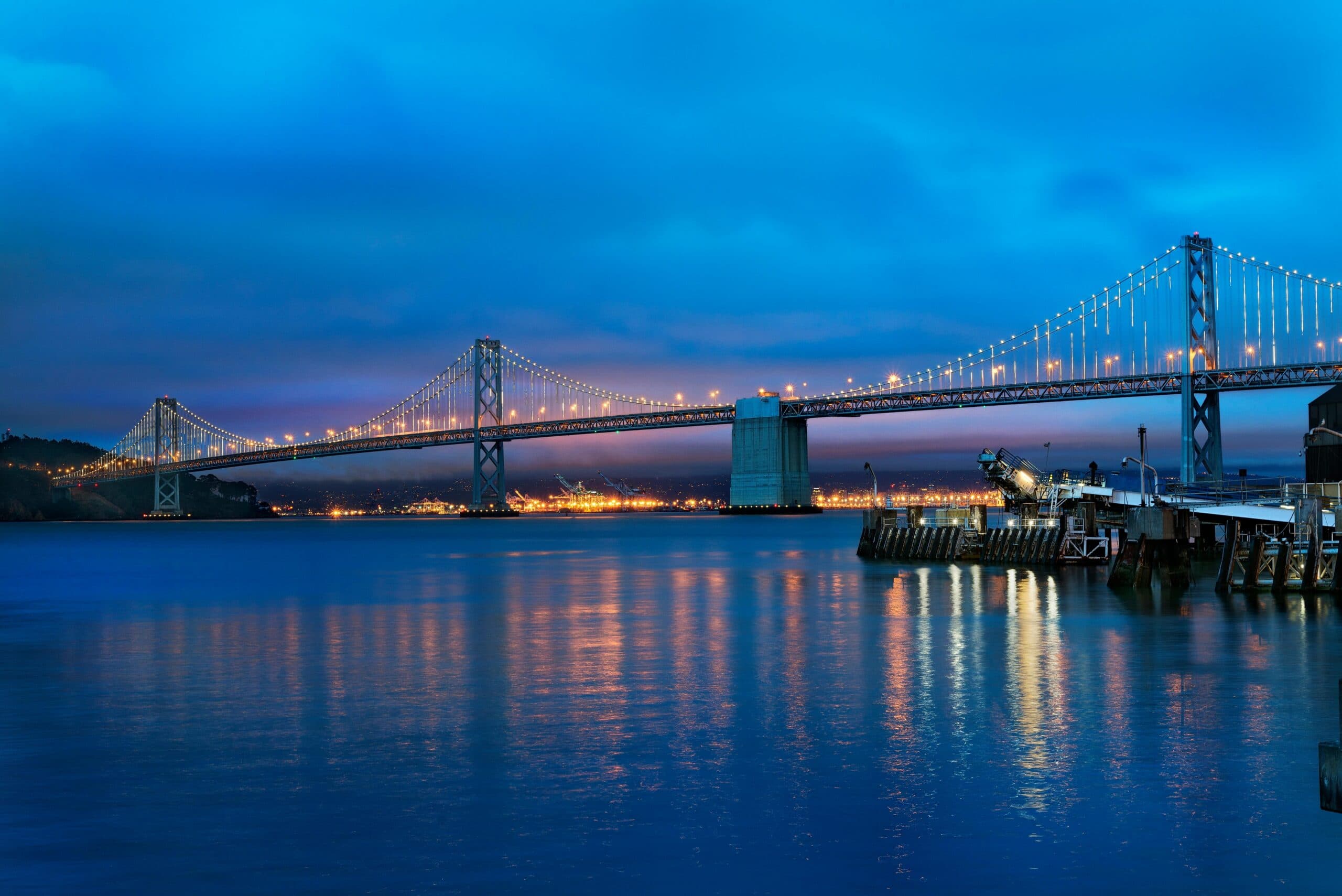 Bay Bridge illuminated at dusk, with its lights reflected across calm bay water and waterfront piers in the foreground.