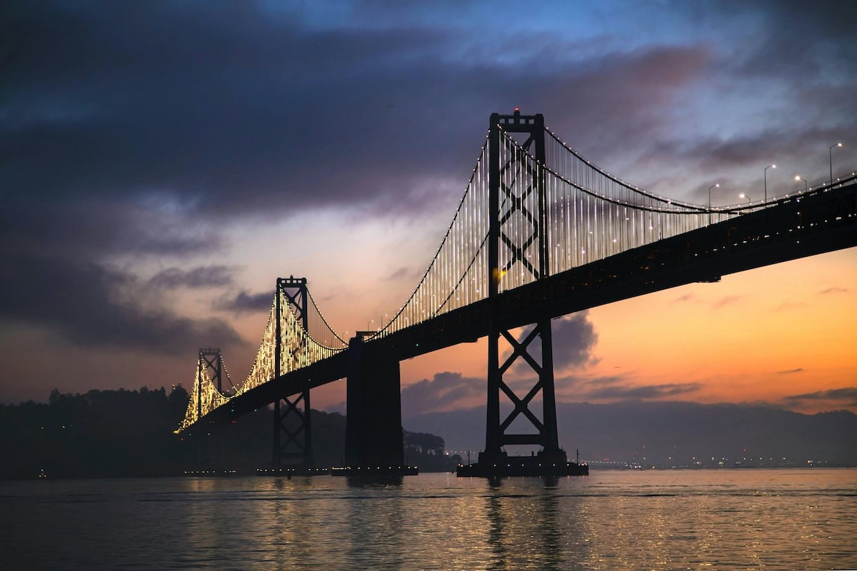 Bay Bridge at sunset, silhouetted against a glowing sky with bridge lights reflecting on the water below.